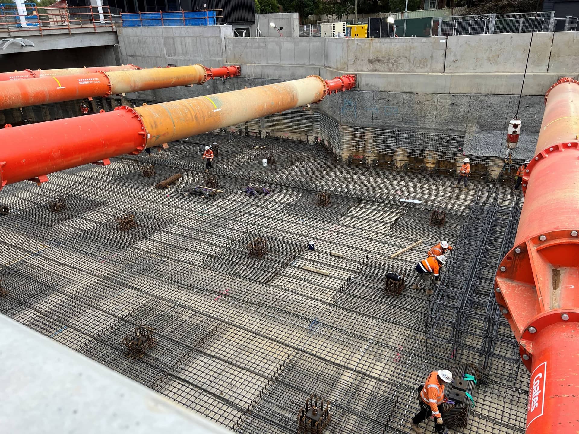 Spartan steel fixers tying a tank slab beneath the orange propping struts of a deep excavation